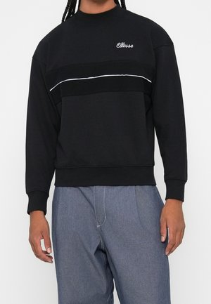 Osoba nosząca czarną bluzę z białym wykończeniem i logo "Ellesse" na piersi, połączoną z luźnymi niebieskimi spodniami.
