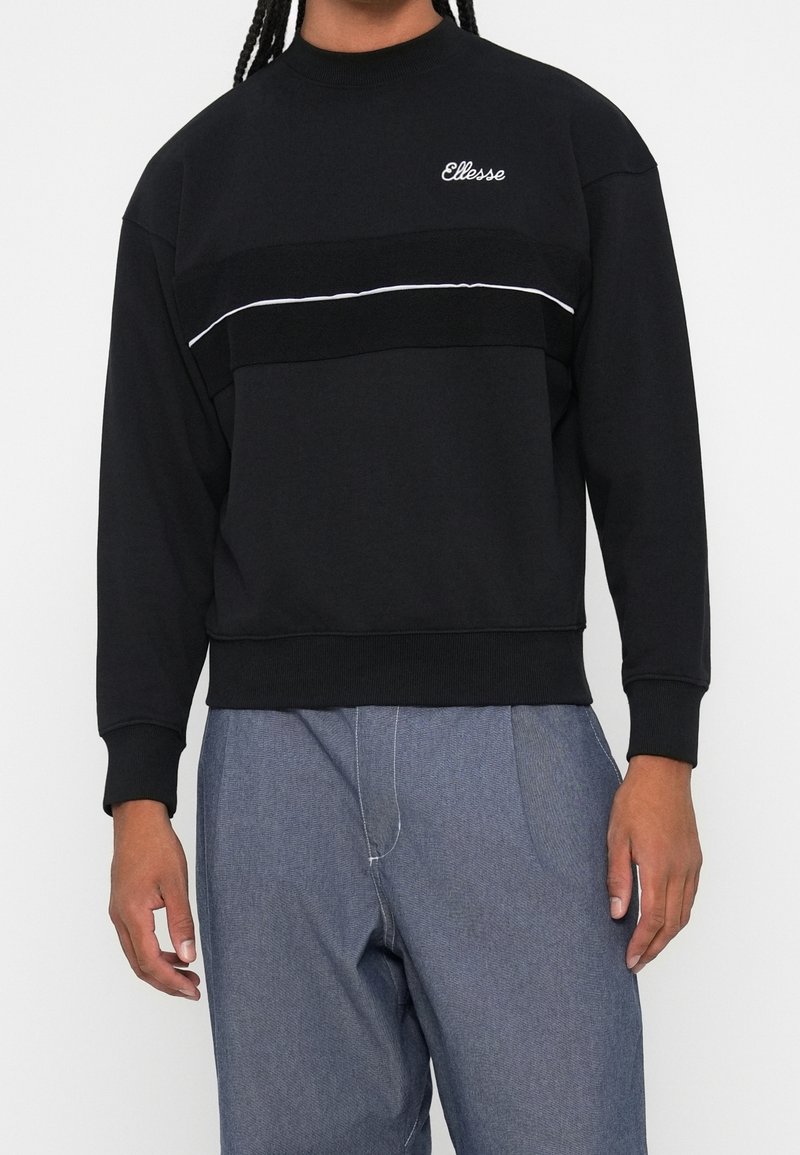 Personne portant un sweat-shirt noir avec des passepoils blancs et le logo "Ellesse" sur la poitrine, associé à un pantalon bleu large.