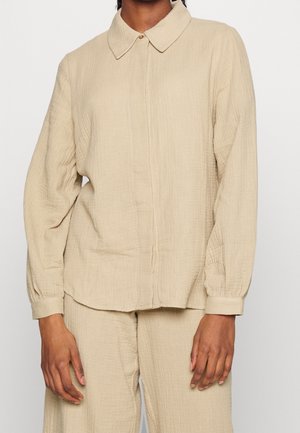 Beige, langärmeliges Hemd mit Knöpfen, Kragen und passenden, lockeren Hosen, getragen von einer stehenden Person mit entspannten Händen.