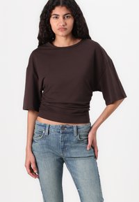 Brown cropped top with short sleeves, featuring a round neckline and gathered detail at the waist, paired with faded blue jeans.
