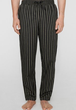 Hombre con pantalones negros con rayas verticales beige y una cintura elástica con cordón, de pie descalzo contra un fondo liso.
