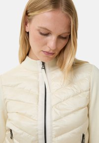 Light beige puffer jacket with a zip front, ribbed collar, and angled quilted design. Features two side zip pockets and contrasting textures.