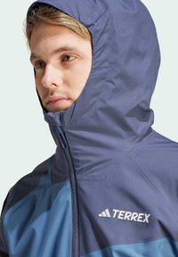 Chaqueta Terrex en azul marino y azul, con un alto cuello y capucha, que presenta un cierre frontal y el logo de la marca en la parte izquierda del pecho. Textura suave.