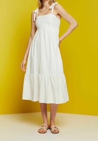 Femme portant une robe mi-longue blanche sans manches avec des bretelles nouées aux épaules et des sandales blanches, debout devant un fond jaune.