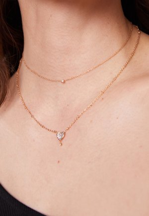 Twee delicate gouden kettingen om de hals van een vrouw, één met een kleine edelsteen en de andere met een hartvormige hanger.