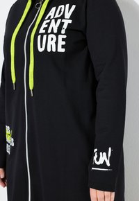 Black zip-up hoodie with neon green drawstrings, white block letters reading "ADVENTURE" on chest, and white stylized letters on left sleeve cuff.