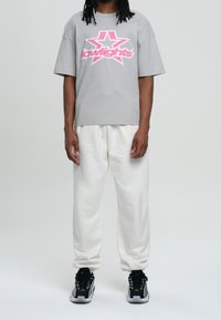 Camiseta gris con logo gráfico de estrella rosa, combinada con pantalones de chándal blancos y zapatillas negras. Diseño casual y con ajuste relajado.