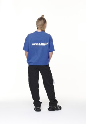 Sininen puuvillapaita, jossa on valkoinen logo selässä; yhdistetty mustiin cargo-housuihin, joissa on sivutaskut, ja mustiin lenkkareihin.