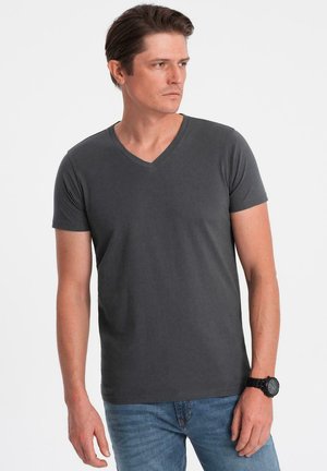 Hombre vestido con camiseta gris oscuro de cuello en V y jeans azules, mirando a la derecha, con un reloj negro en la muñeca izquierda, frente a un fondo blanco.