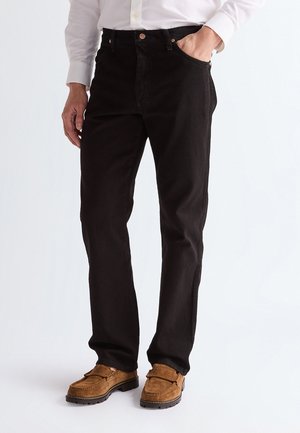 Muž nosící černé džíny s rovnými nohavicemi, bílou košili na knoflíky a hnědé semišové mokasíny, stojící s jednou rukou v kapse.