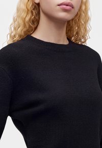 Personne blonde aux cheveux bouclés portant un pull en laine noir uni, montrant le cou et le haut de la poitrine sur un fond neutre.