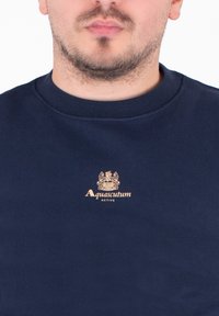Granatowy sweter z okrągłym dekoltem, wykonany z miękkiego materiału. Posiada złotą haftowaną naszywkę "Aquascutum ACTIVE" na klatce piersiowej.