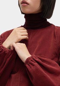 Bordowy top z korduroyu z wysokim kołnierzem, z obszernymi, marszczonymi rękawami i teksturowanym wykończeniem. Zbliżenie na dłonie umieszczone przy kołnierzu.