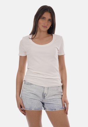 Frau mit glatten braunen Haaren, die ein weißes kurzärmeliges Oberteil mit Rundhalsausschnitt und hellblaue Jeans-Shorts trägt, steht in entspannter Haltung.