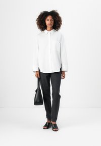Camisa blanca de manga larga con botones y bolsillo, combinada con jeans gris oscuro y sandalias negras sin cordones; el modelo lleva un bolso negro.