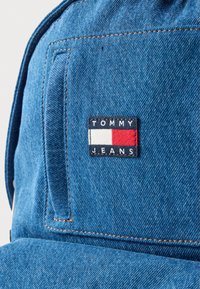 Modrá rifľová čiapka s textúrovaným povrchom. Obsahuje obdĺžnikovú vyšívanú nášivku s nápisom "TOMMY JEANS" a farebnou kombináciou bielej, červenej a námorníckej modrej.