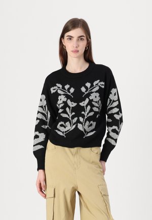 Sort strikket trøje med et blomsterprint i grå, rund halsudskæring og løs pasform. Båret over beige cargo bukser.