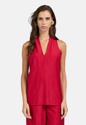 Donna con lunghi capelli castani che indossa grandi orecchini di perle e un top rosso senza maniche con scollo a V e pantaloni rossi abbinati.