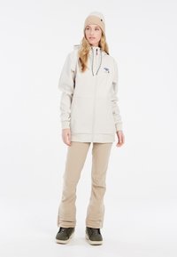 Krem kaput s kapuljačom, prednjim patentnim zatvaračem i šarenim logotipom. Bež hlače, teksturirani materijal. Crne i zelene cipele.