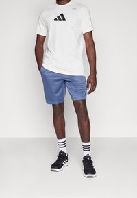 Bílý tričko s krátkým rukávem a černým logem Adidas, v kombinaci se světle modrými šortkami, černými teniskami a bílými ponožkami s černými pruhy.