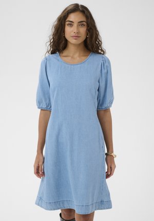 Femme aux cheveux bouclés portant une robe en denim bleu clair avec des manches courtes bouffantes et un col rond, sur un fond uni.