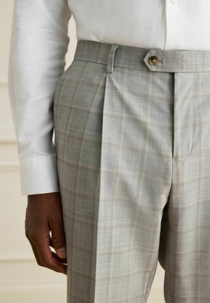 Man wearing light gray plaid tailored trousers with a pleat and tab closure, paired with a white long-sleeve shirt.