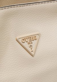 Beżowa skórzana torebka z trójkątną złotą zawieszką z logo z napisem „GUESS USA”, o fakturowanej powierzchni i gładkim przeszyciu krawędzi.