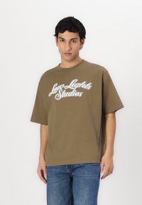 Oliven grøn T-shirt med korte ærmer og rund hals, med hvidt grafisk tekst "Low Lights Studios" på forsiden. Blødt bomuldsstof.