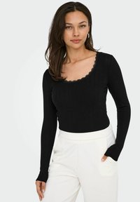 Schwarzes, geripptes Langarmoberteil mit einem gewellten Ausschnitt. Kombiniert mit hoch taillierten weißen Hosen, die eine schlanke Silhouette betonen.
