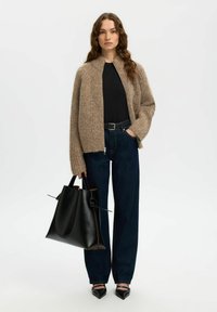 Cardigan en tricot beige avec un col haut, associé à une chemise noire et un jean foncé taille haute. Sac fourre-tout en cuir noir et chaussures pointues.