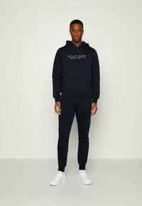 Hanorac bleumarin cu logo "HILFIGER NEW YORK" în alb și roșu, asortat cu pantaloni de trening bleumarin și adidași albi. Material neted, croială lejeră.