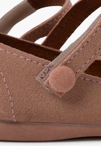 Zapato de ante marrón con acabado texturizado, correa ajustable con cierre de botón redondo y suave, y acentos de costura en contraste.