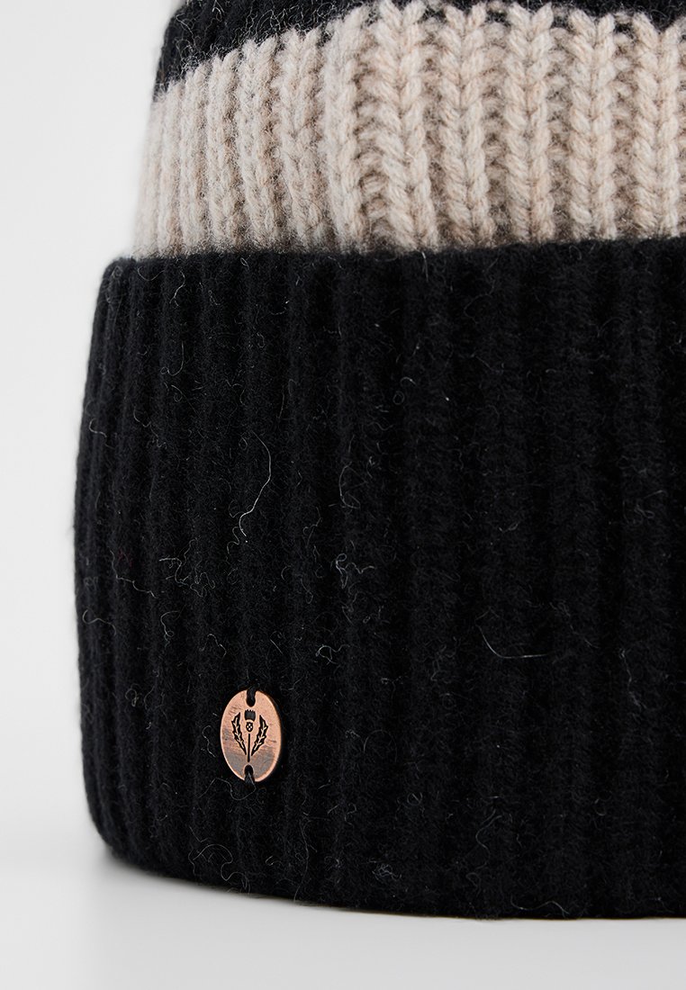 Dwukolorowa czapka typu beanie z prążkowanego materiału, z jasnobeżową górną częścią, czarną dolną częścią oraz małym miedzianym emblematem z wzorem ostrokrzewu na mankiecie.