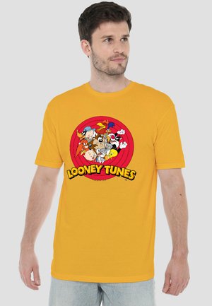 Mann trägt ein gelbes T-Shirt mit einem roten, runden Looney Tunes-Logo mit mehreren Cartoon-Figuren in der Mitte.