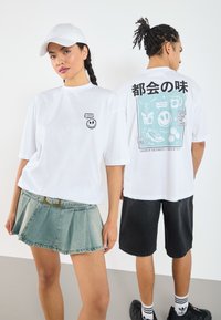 Camiseta blanca de algodón con un gráfico de una cara sonriente en el pecho y coloridas ilustraciones de comida en la parte posterior, combinada con una falda de mezclilla.