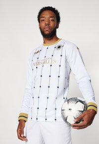 Pitkähiha valkoinen jalkapollopaita, jossa on sininen timanttikuosi ja kultaiset yksityiskohdat. Sisältää "Football Club de Versailles" -brändäyksen. Pitelee kuviollista jalkapalloa.