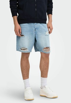 Hombre con pantalones cortos vaqueros rotos de color azul claro, calcetines blancos, zapatillas blancas y una sudadera negra con cremallera, de pie sobre un fondo liso.