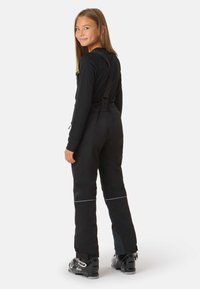 Musta hiihtohaalari, jossa on säädettävät olkaimet ja takataskut, heijastavia yksityiskohtia sekä istuva malli, yhdistettynä hiihtokenkiin.