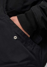 Zwarte jas met een ritszak, voorzien van een ribbelmanchet en een zichtbare zilveren drukknoop aan de zijkant, met een gladde, matte textuur.