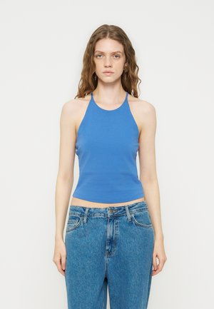 Νέα γυναίκα με μακριά σγουρά μαλλιά, που φοράει μπλε αμάνικο τοπ με λαιμόκοψη και μπλε ψηλόμεσα τζιν, στέκεται μπροστά σε μονόχρωμο φόντο.