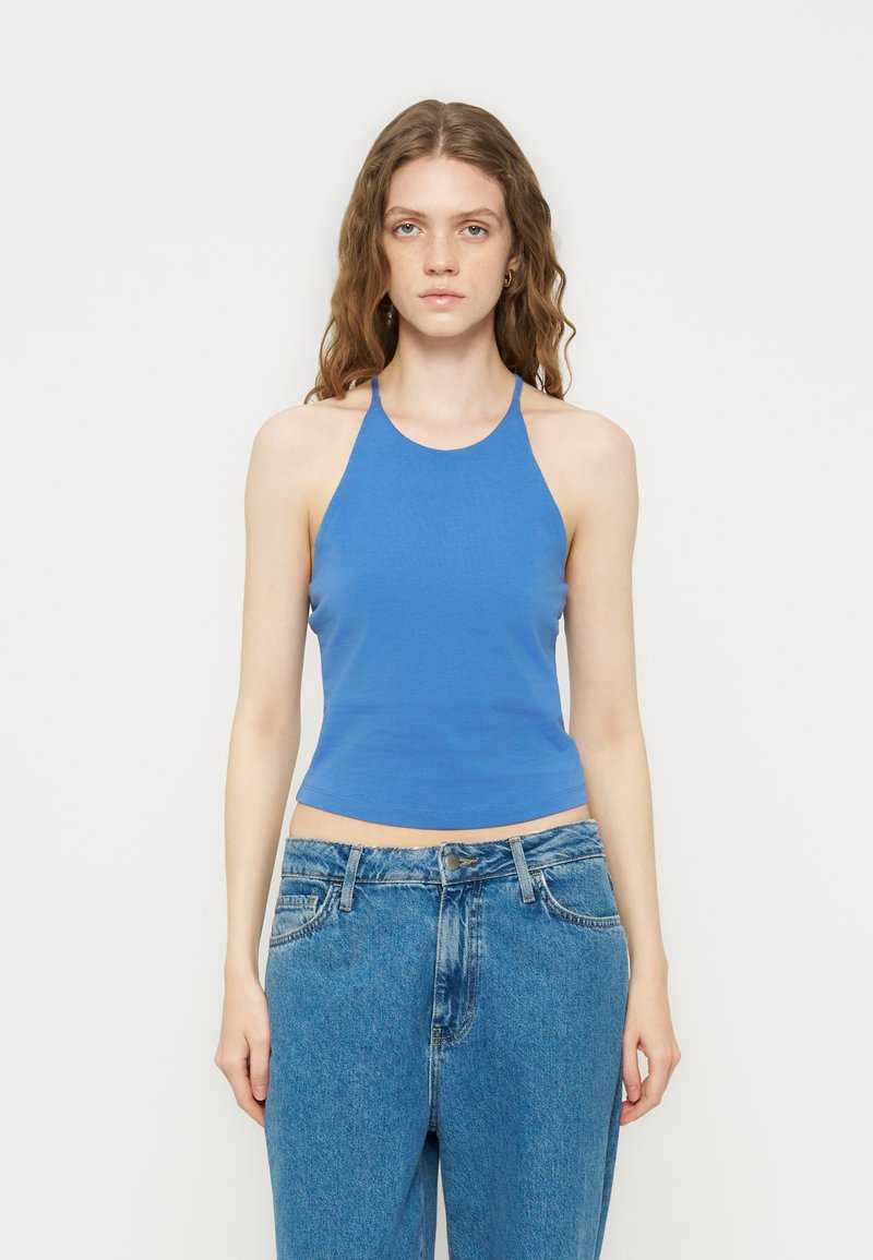 Νέα γυναίκα με μακριά σγουρά μαλλιά, που φοράει μπλε αμάνικο τοπ με λαιμόκοψη και μπλε ψηλόμεσα τζιν, στέκεται μπροστά σε μονόχρωμο φόντο.