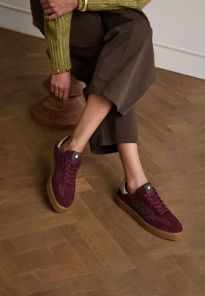 Bordeaux suède sneakers met textuurdetails, leren accenten en een dikke gumzool, opgesteld op een gepolijste houten vloer.