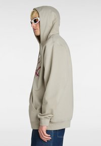 Beige hoodie met een voorzak, voorzien van een roze grafisch ontwerp op de borst. Gestyled met blauwe jeans en witte zonnebril.