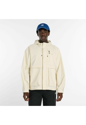 Giovane uomo che indossa una giacca con cappuccio beige chiaro, pantaloni neri e un berretto da baseball blu, in piedi davanti a uno sfondo bianco uniforme.