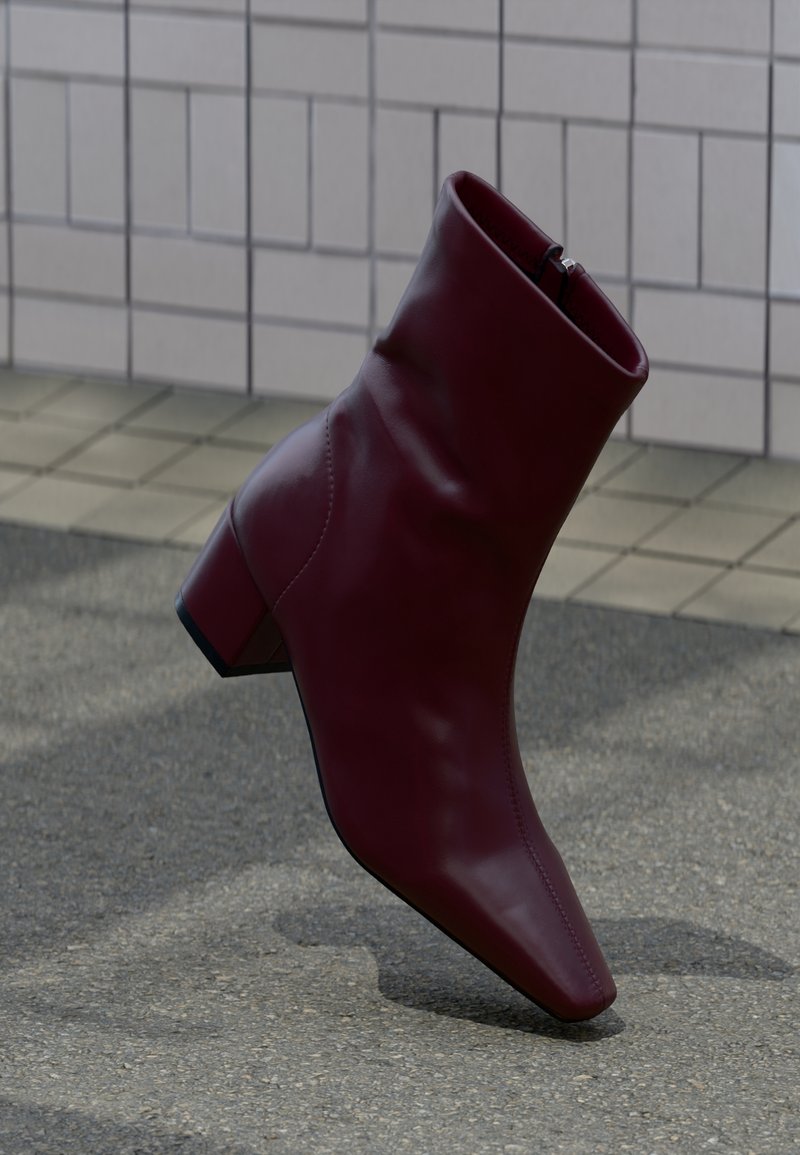 Une botte cheville en cuir bordeaux avec un talon bloc sur une surface pavée grise près de murs carrelés.
