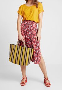 T-shirt gialla a maniche corte abbinata a una maxi gonna rossa a fantasia floreale. Accentata da una borsa a tracolla intrecciata a righe e sandali con tacco rossi.