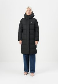 Svart pufferjakke med høy krage og frontglidelås, med kviltede seksjoner. Kombinert med vide denimjeans og lyse sko.