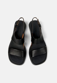 Sandalias de cuero negro con correas anchas, diseño de talón abierto, suela plana y marca Camper grabada en la plantilla. Textura suave en todo su diseño.