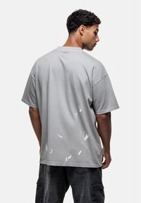 T-shirt en coton gris oversize à manches courtes, présentant des éclaboussures de peinture blanche irrégulières au dos. Encolure simple et coupe décontractée.