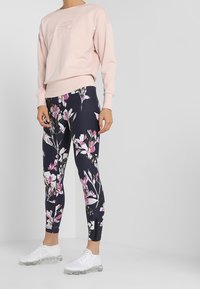 Světle růžová mikina s nápisovým designem, spárovaná s tmavými květinovými legínami s růžovými a bílými květy. Bílé sportovní boty.